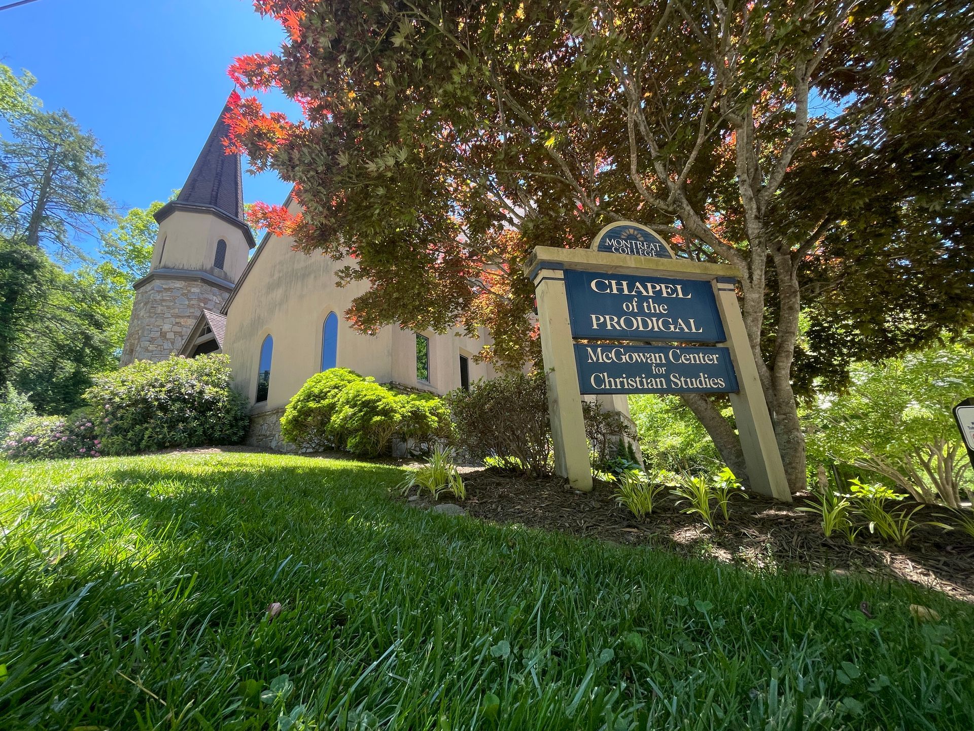 Church with red roof and blue sign on a sunny, tree-shaded hillside