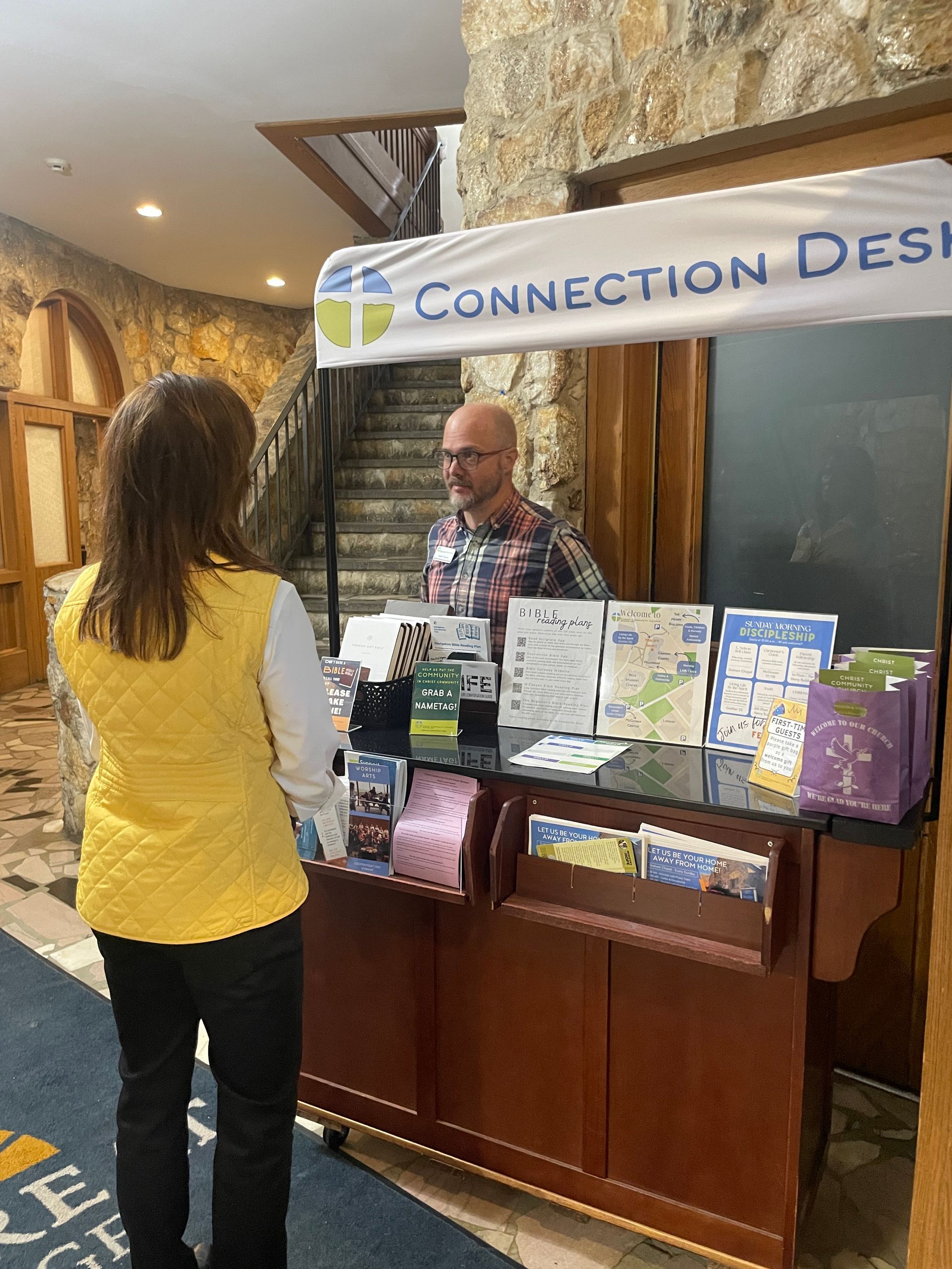 Woman speaking with a man behind a Connection Desk information kiosk in the lobby of a stone building.