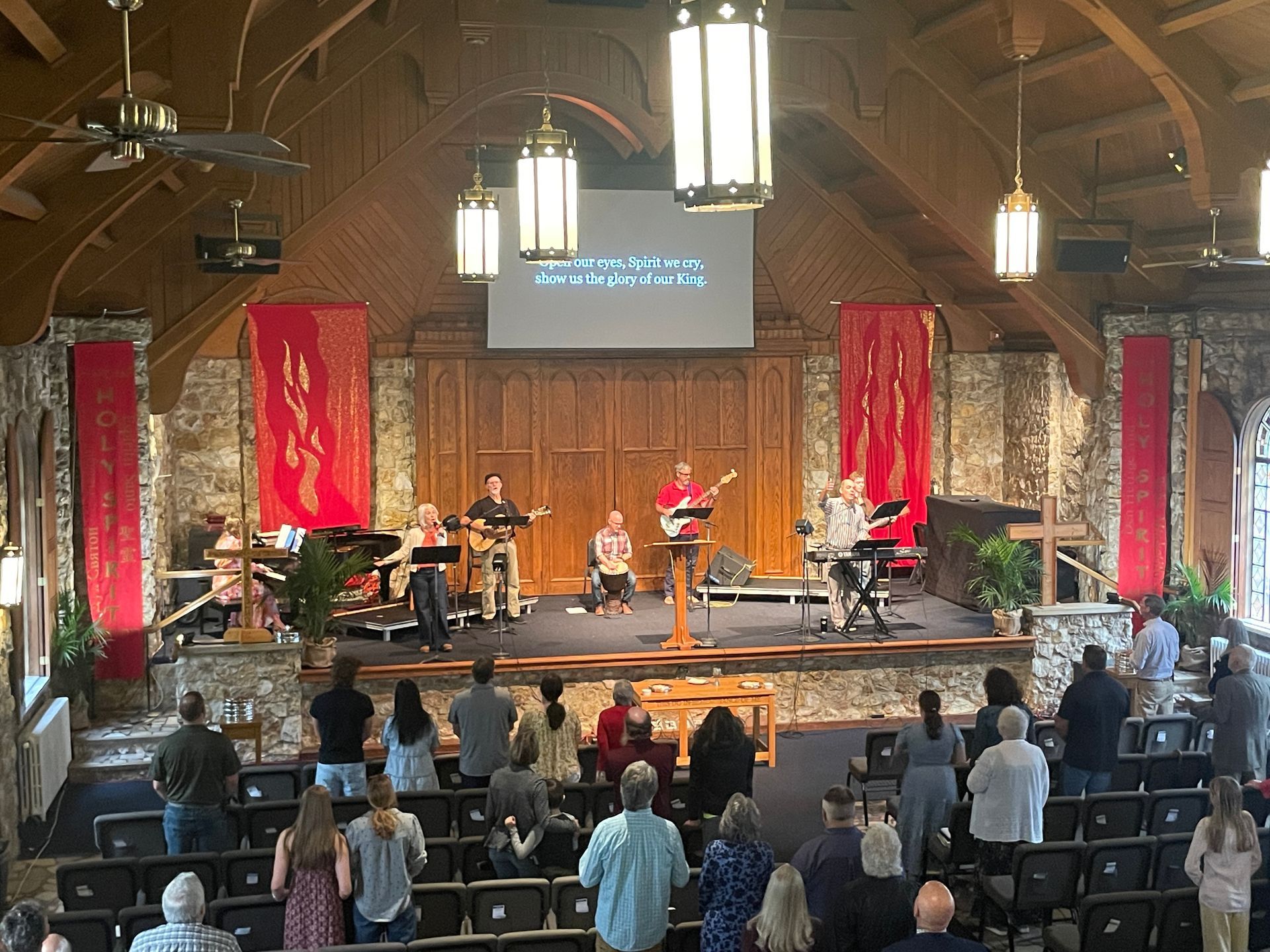 Live band performing on a church-style stage with audience seated in front, under warm hanging lights.
