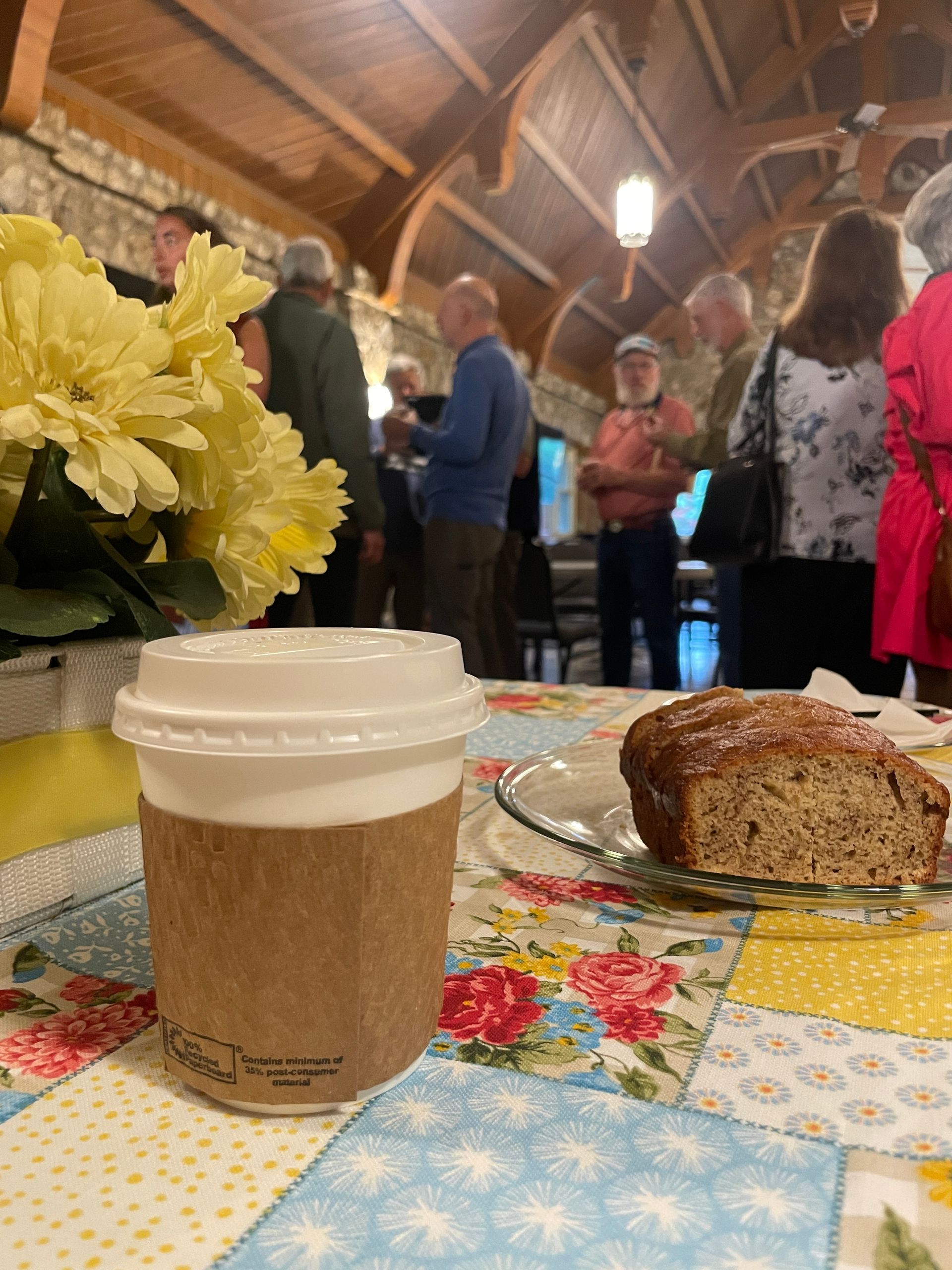 Coffee cup and banana bread on a table with yellow flowers with people mingling and socializing in the background. 