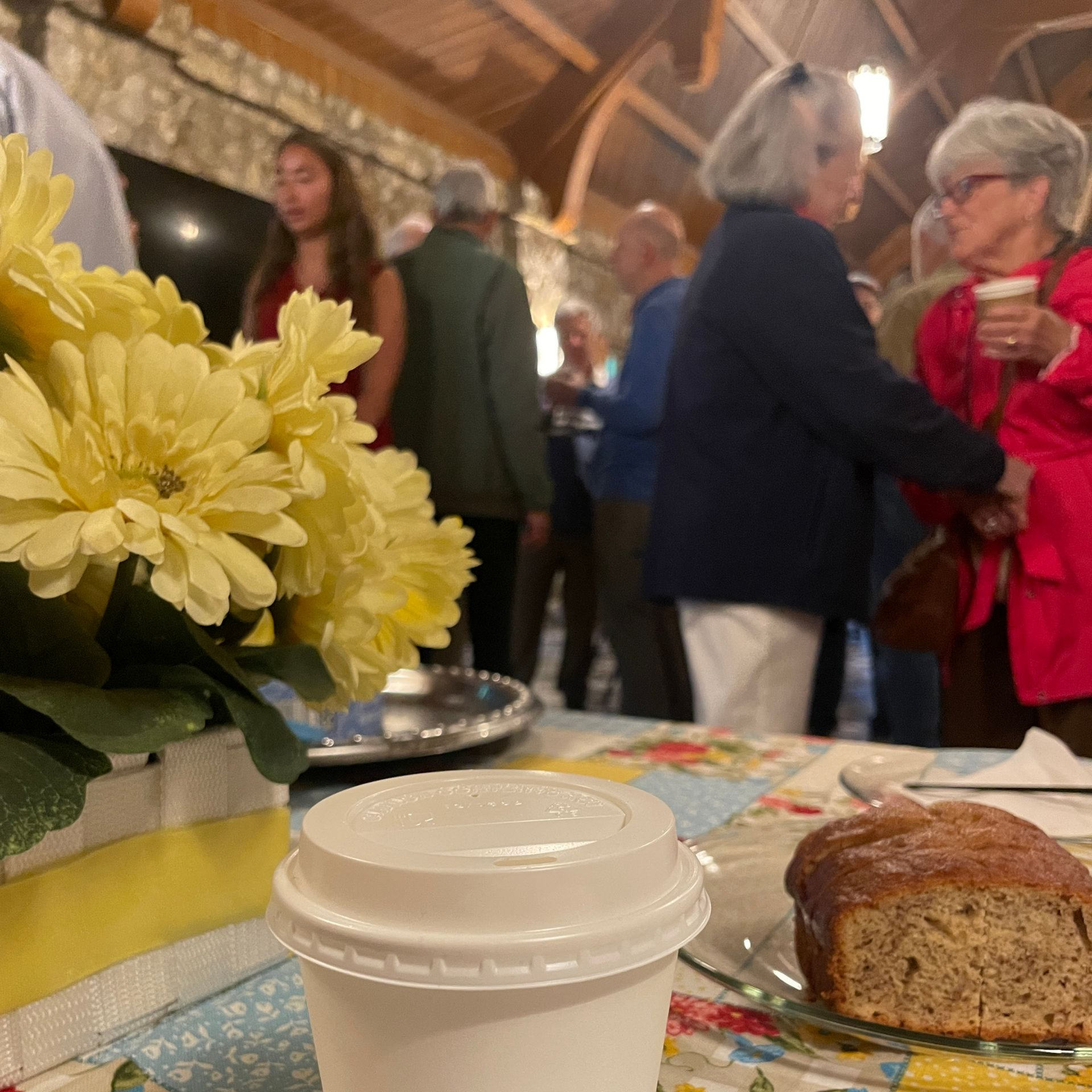 People mingling and talking with a foreground of a serving table with coffee cup, baked goods, and floral arrangement.