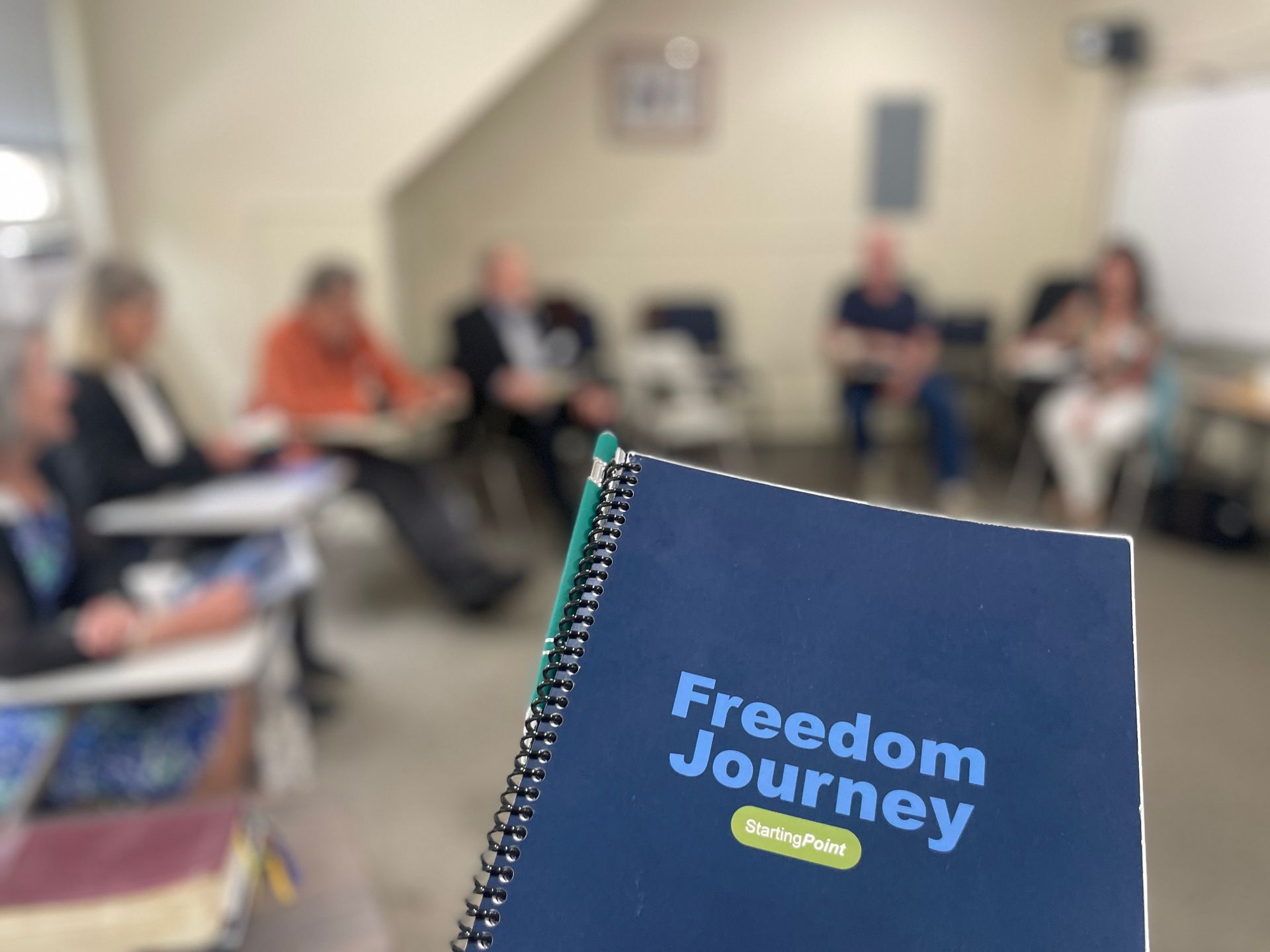 Notebook titled “Freedom Journey” in foreground, with people seated in a blurred meeting room behind it.