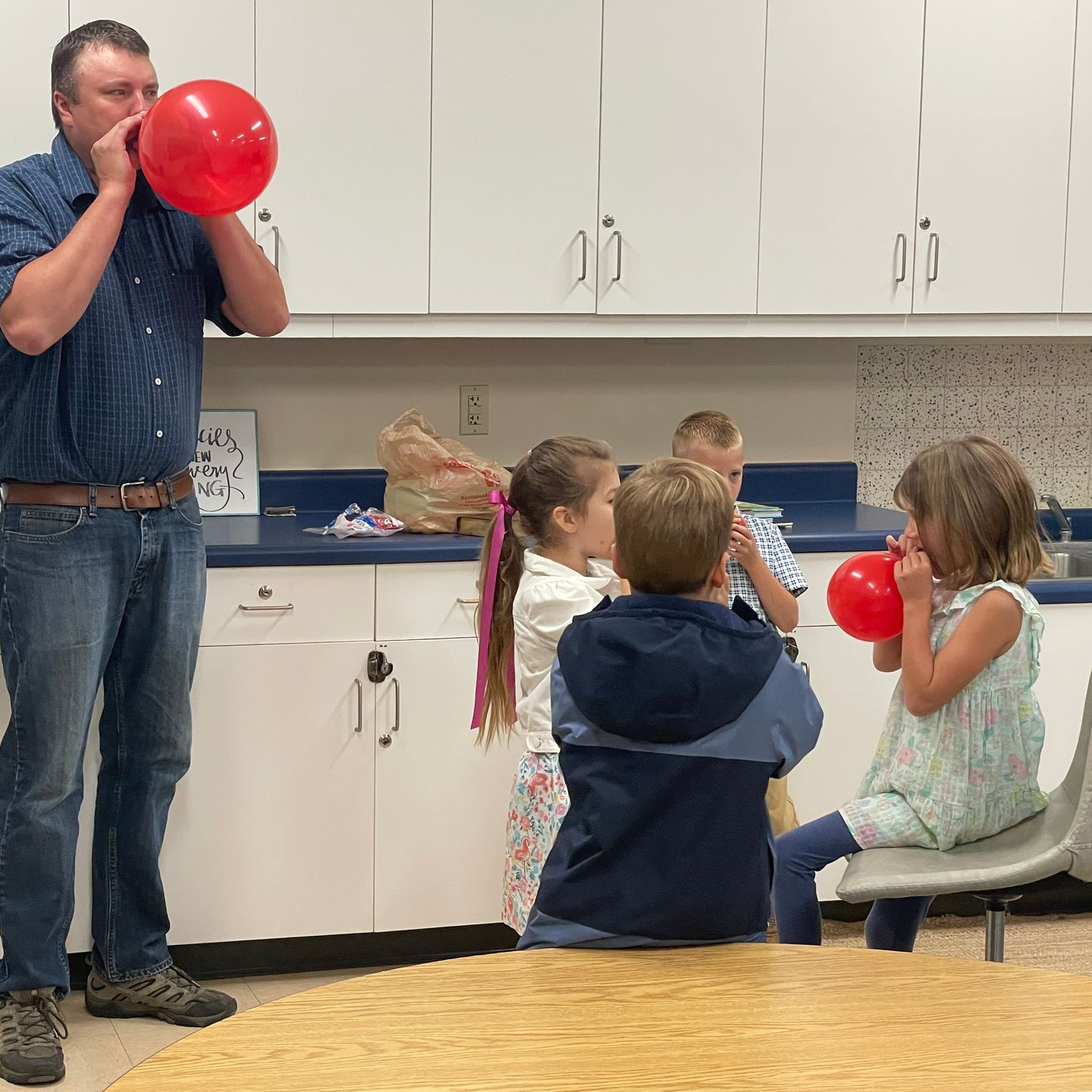Adult demonstrating balloon blowing to children in a classroom with white cabinets.