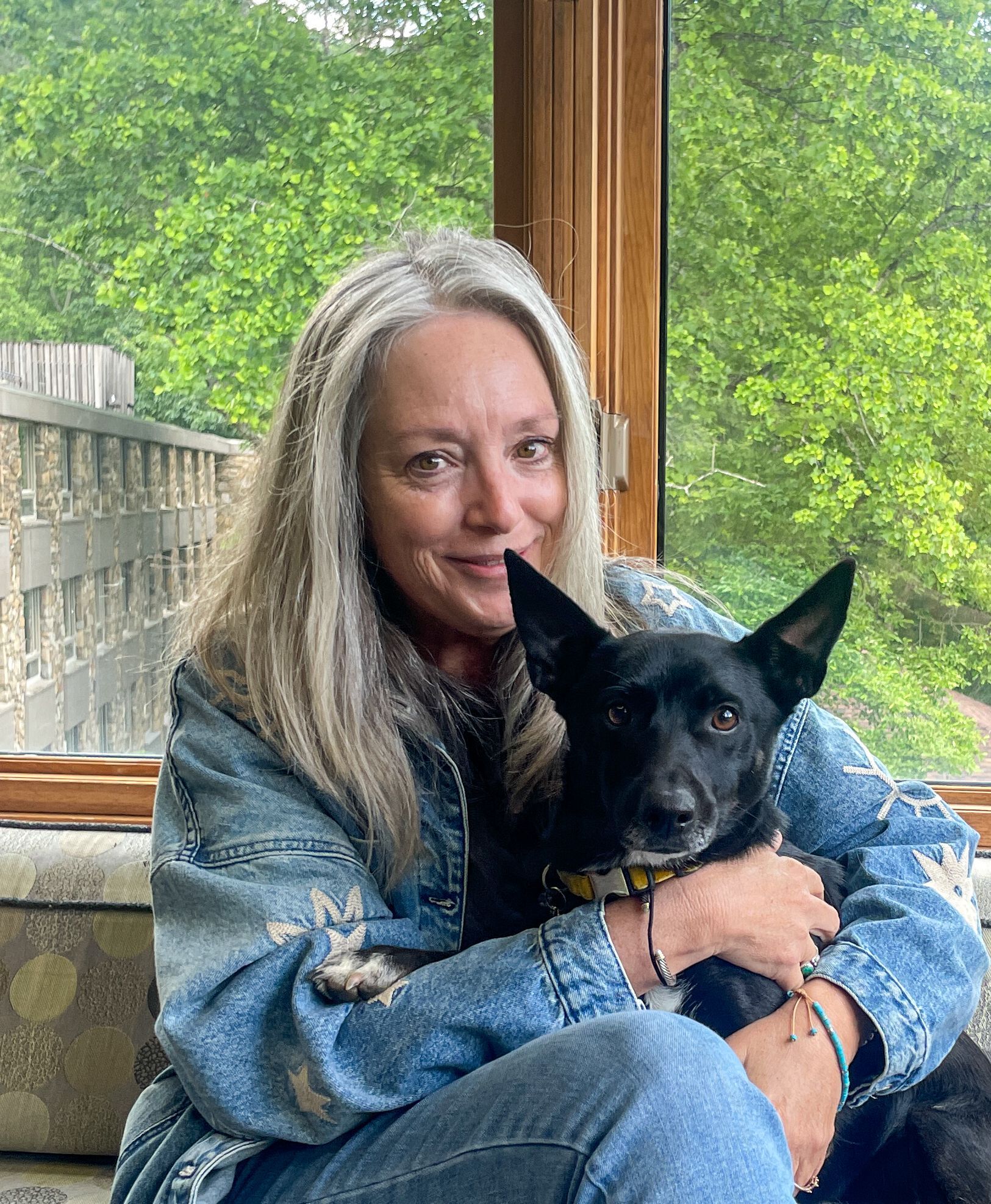 Woman in a jeans jacket hugging a black dog with greenery outside the window behind.