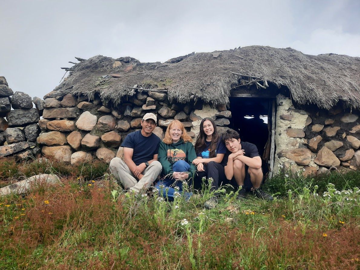 Family of four sitting in front of a stone hut with thatched roof.