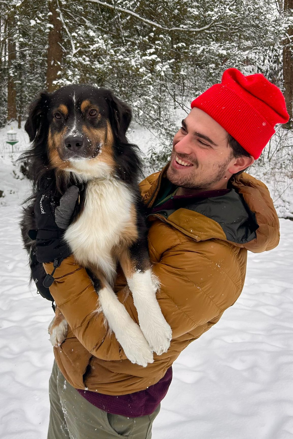 Man in a red hat holding a black, white, and brown dog, standing in the snow.