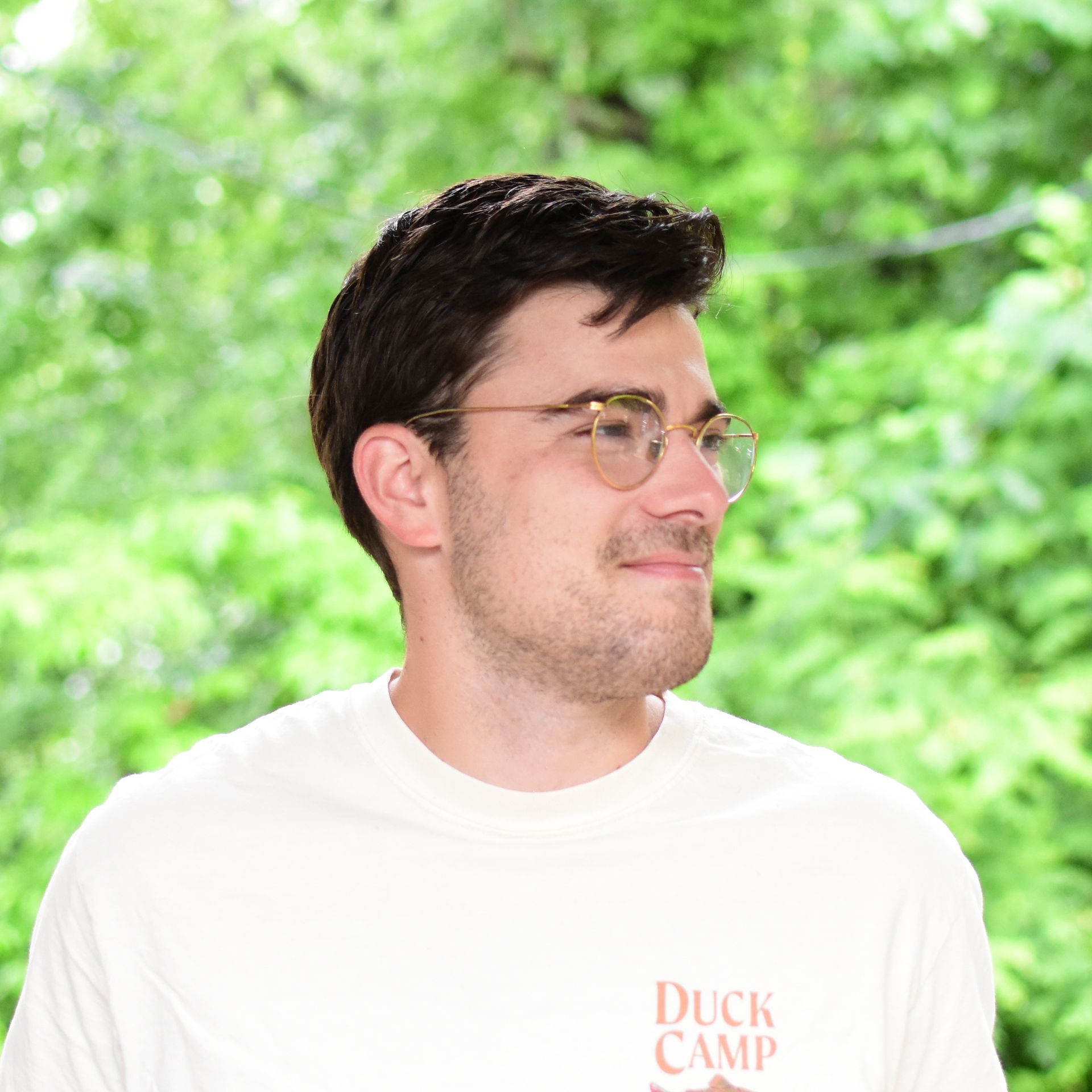 Man wearing glasses and a white Duck Camp shirt, looking to the side outdoors against a green background
