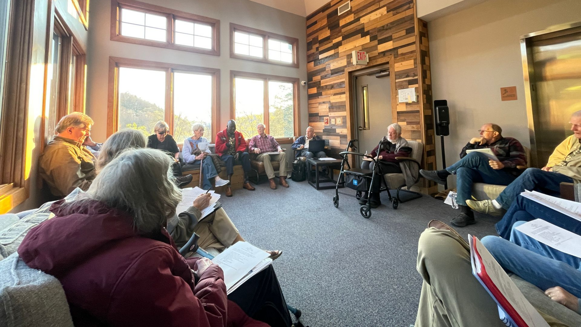 People seated in a sunlit living room for a group discussion, with large windows and a wood plank accent wall.