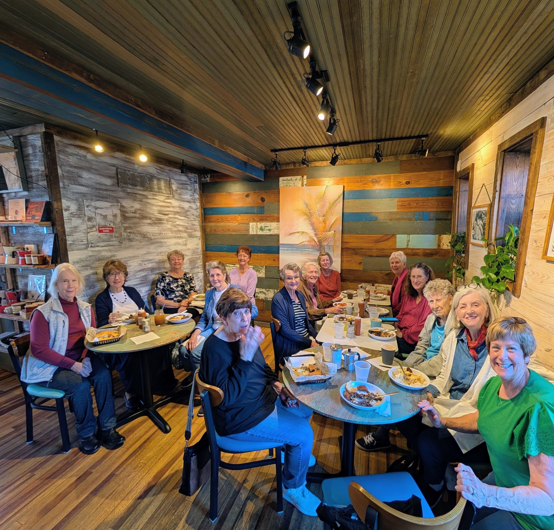 Group of people seated around tables in a cozy rustic café, smiling and chatting over drinks and food.