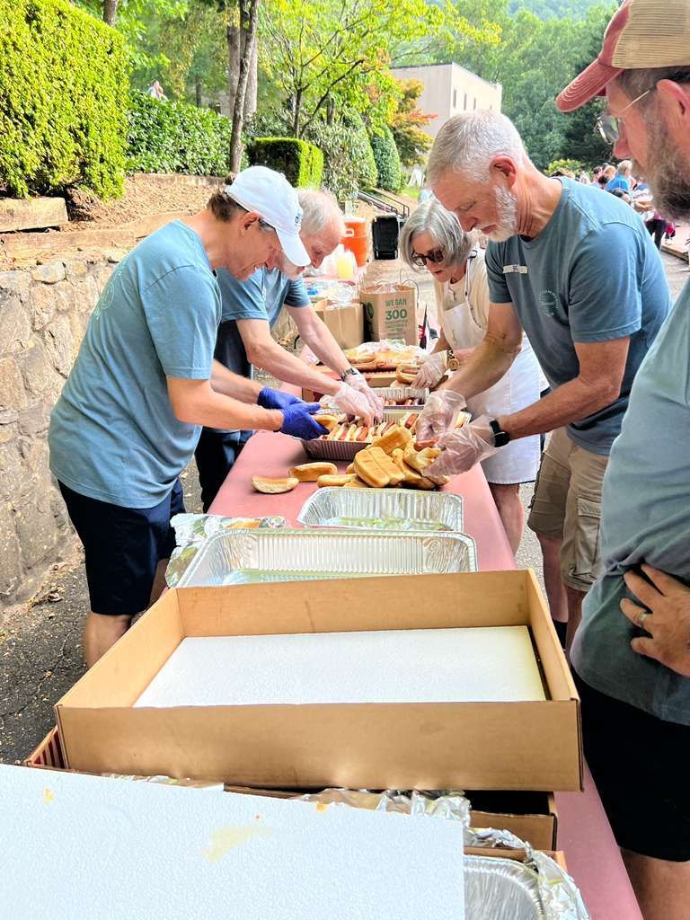 People serving food at a long outdoor table with trays and containers during a community meal