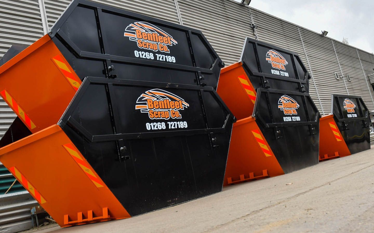 Four black and orange skips with Benfleet Scrap logos, lined up against a building.