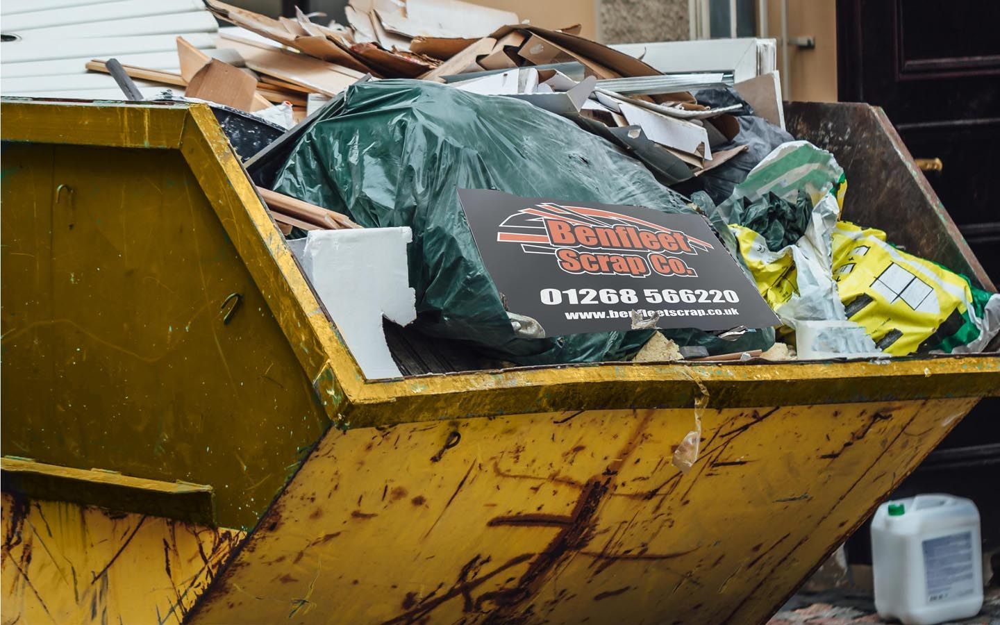 A yellow skip filled with rubbish and a sign that says 'Benfleet Scrap ' on it.