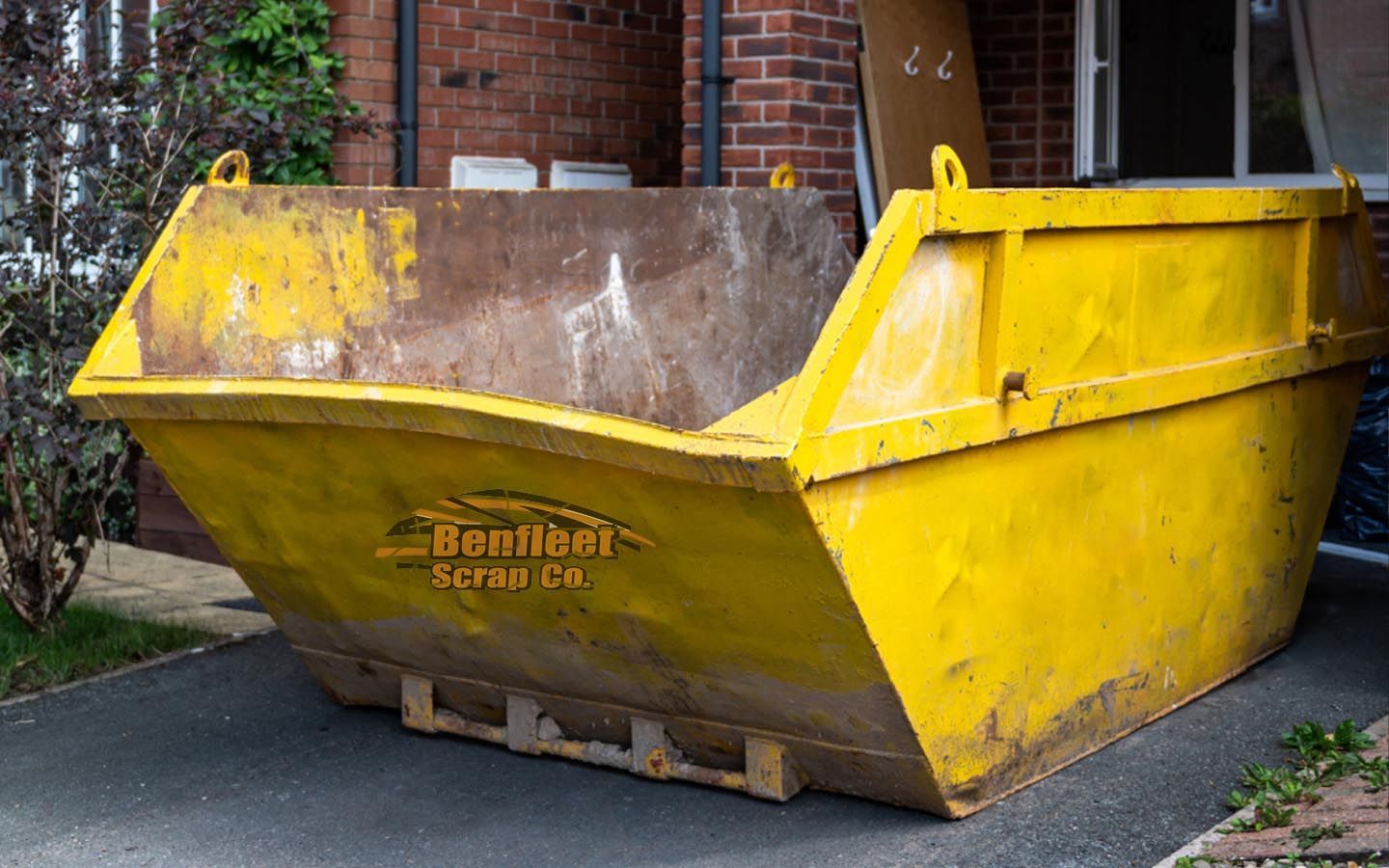 A yellow skip is parked in front of a brick building with Benfleet Scrap logo