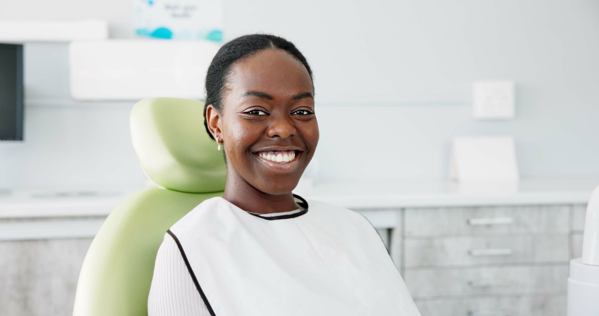 Smiling person in a dental chair wearing a bib; bright office setting.