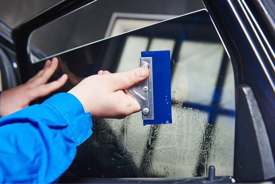 Hand using a blue squeegee to clean a car window with water droplets