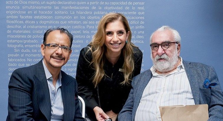 Tres personas posando juntas en el interior, sonriendo frente a una pared azul cubierta de texto.