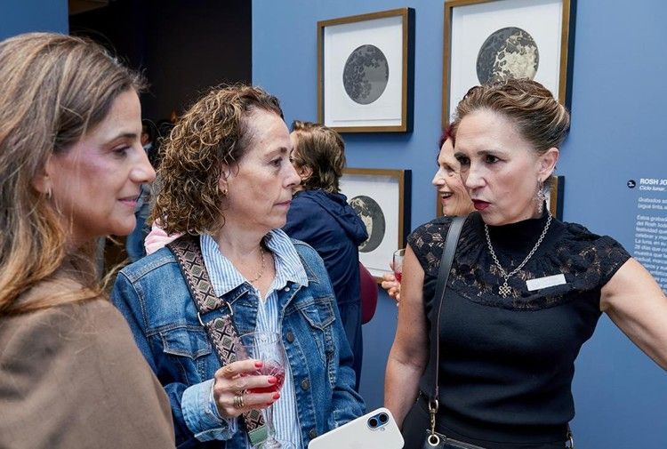 Tres personas conversando en una galería, una de ellas sosteniendo una copa de vino, con obras de arte enmarcadas en paredes azules.