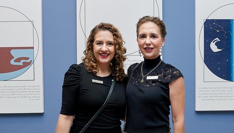Dos mujeres con vestidos negros de pie juntas frente a un cuadro enmarcado, sonriendo a la cámara.