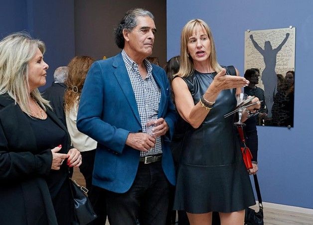 Tres personas se encuentran en una galería, una mujer gesticulando mientras observan las obras de arte en la pared.