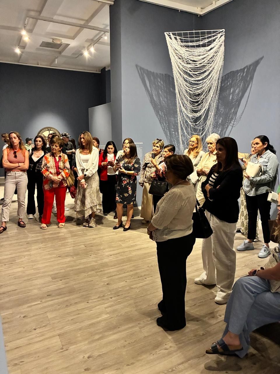 Un grupo de personas se reunió en una galería para escuchar a un orador frente a una obra de arte textil colgada en la pared.