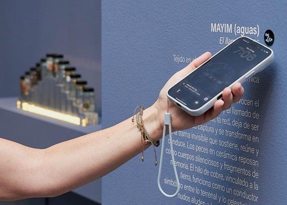 Persona sosteniendo un teléfono inteligente sobre una pantalla digital montada en la pared en un entorno similar a un museo.