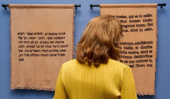 Una persona con un suéter amarillo lee dos pergaminos colgantes con texto en una pared azul.