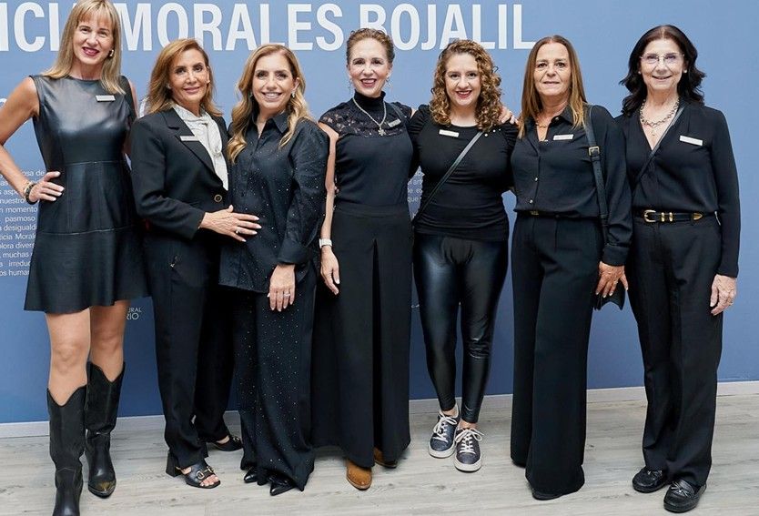 Seis mujeres vestidas de negro posando frente a un escenario para un evento formal.