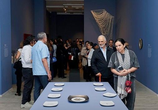 Los visitantes en una galería de paredes azules observan pequeños platos sobre una mesa con una escultura mural en forma de red detrás de ellos.