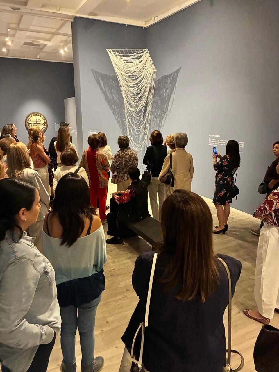 Los visitantes de la galería pueden contemplar una gran escultura mural tejida en blanco, ubicada en un espacio de exposición con paredes azules.