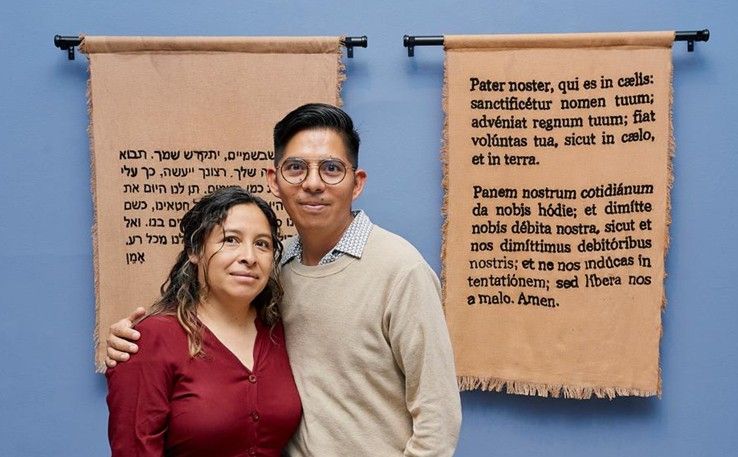 Dos personas de pie frente a pergaminos colgados en la pared con texto manuscrito, uno azul y otro color canela.