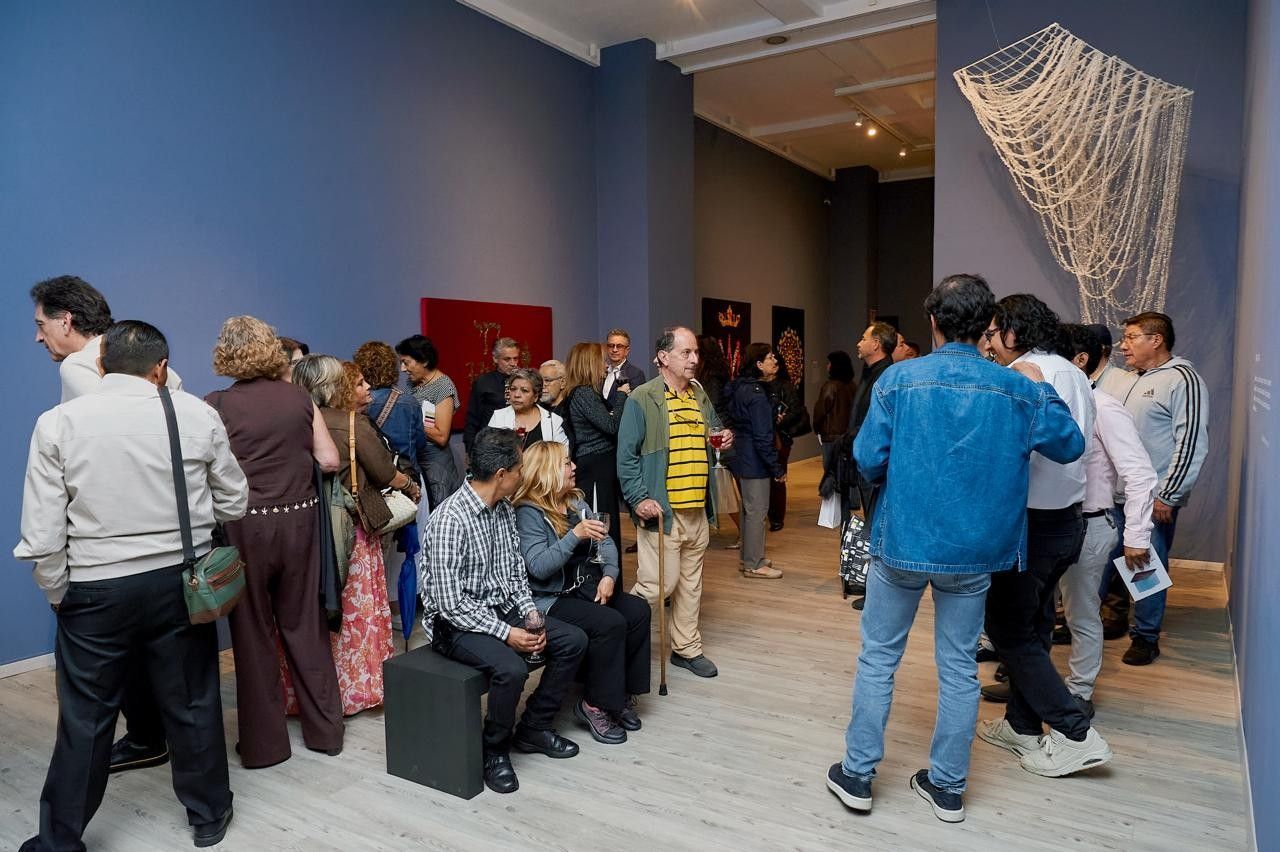 Galería abarrotada de gente charlando, espectadores sentados y grandes obras de arte colgadas sobre paredes azules.