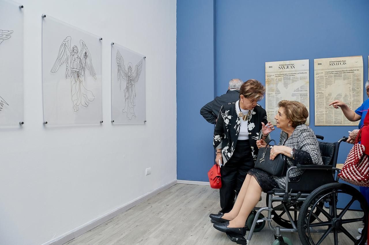 Personas en sillas de ruedas contemplando obras de arte en una galería con paredes azules y cuadros blancos.