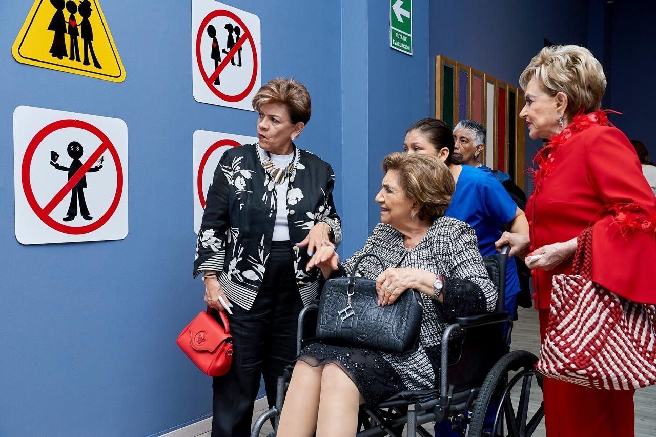 Varias personas se encuentran frente a un edificio azul con letreros de advertencia; una persona está sentada en una silla de ruedas mientras otras observan.