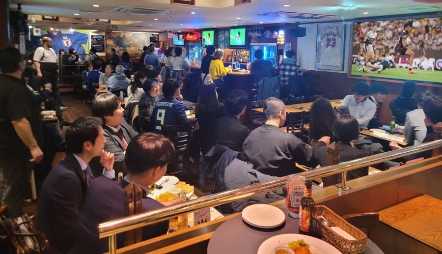 A crowd of people are sitting at tables in a restaurant watching a rugby game.
