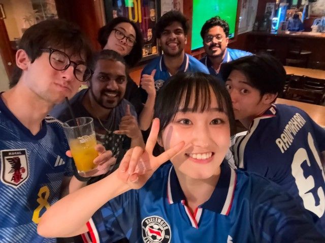 Group selfie of six people smiling in blue jerseys, one holding a drink and another flashing a peace sign.