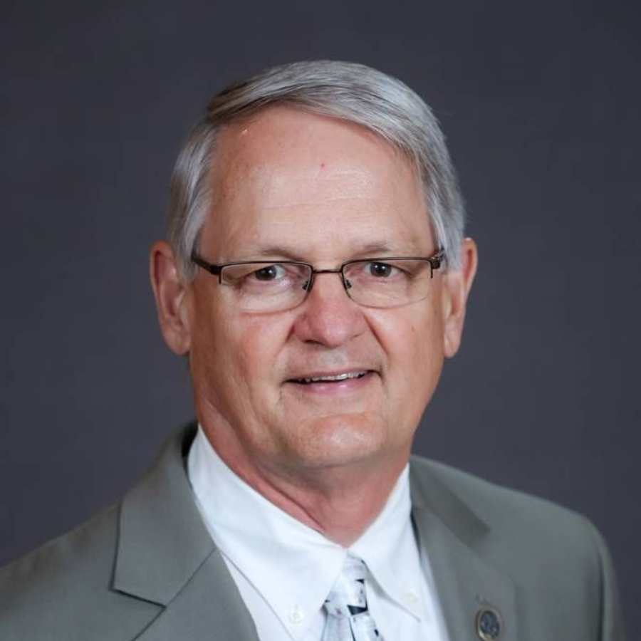 A person with gray hair and glasses wearing a light green suit, white shirt, and patterned tie, against a dark background.
