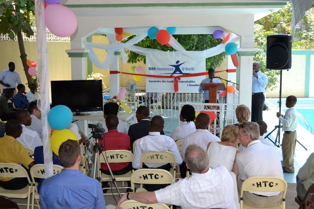 A crowd sits outdoors at an event, facing a speaker at a podium in front of a banner under a decorated trellis structure.