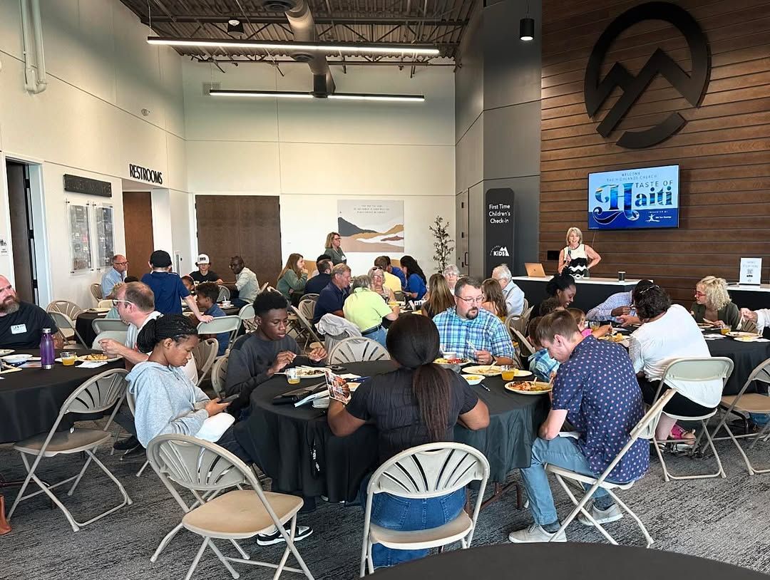 People gather at round tables in a modern, high-ceilinged room with a wooden accent wall and a large company logo.