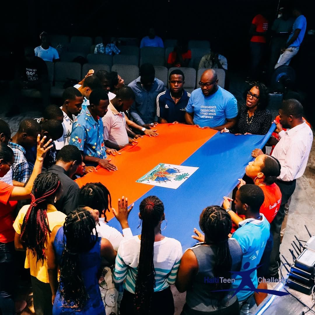 A diverse group of people surrounds and touches a large blue and orange flag with a central emblem in a dark hall.