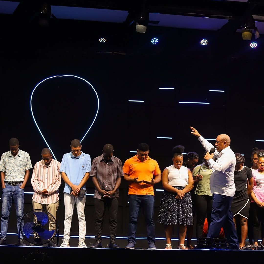 A person leads a group standing on a stage in front of a dark backdrop with illuminated light patterns.
