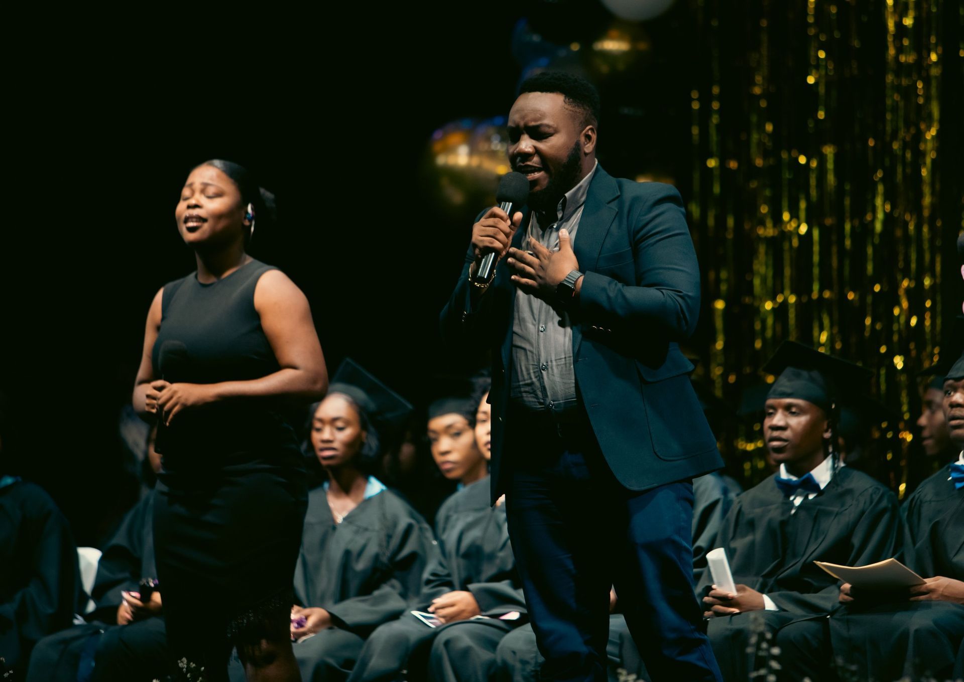 Two individuals perform on stage with microphones in front of a seated audience dressed in graduation attire.