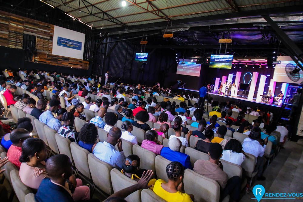 A large, diverse audience sits in a dimly lit auditorium facing a stage with bright displays and musicians.