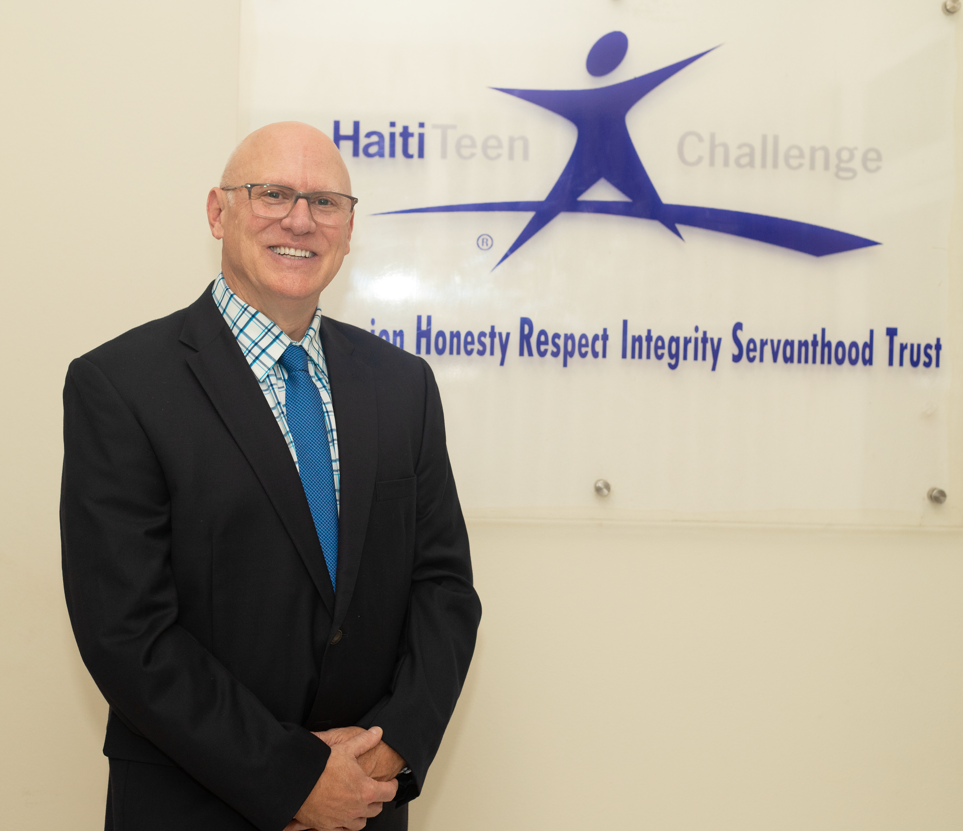 A person in a suit and blue tie stands before a Haiti Teen Challenge sign listing values like honesty and integrity.