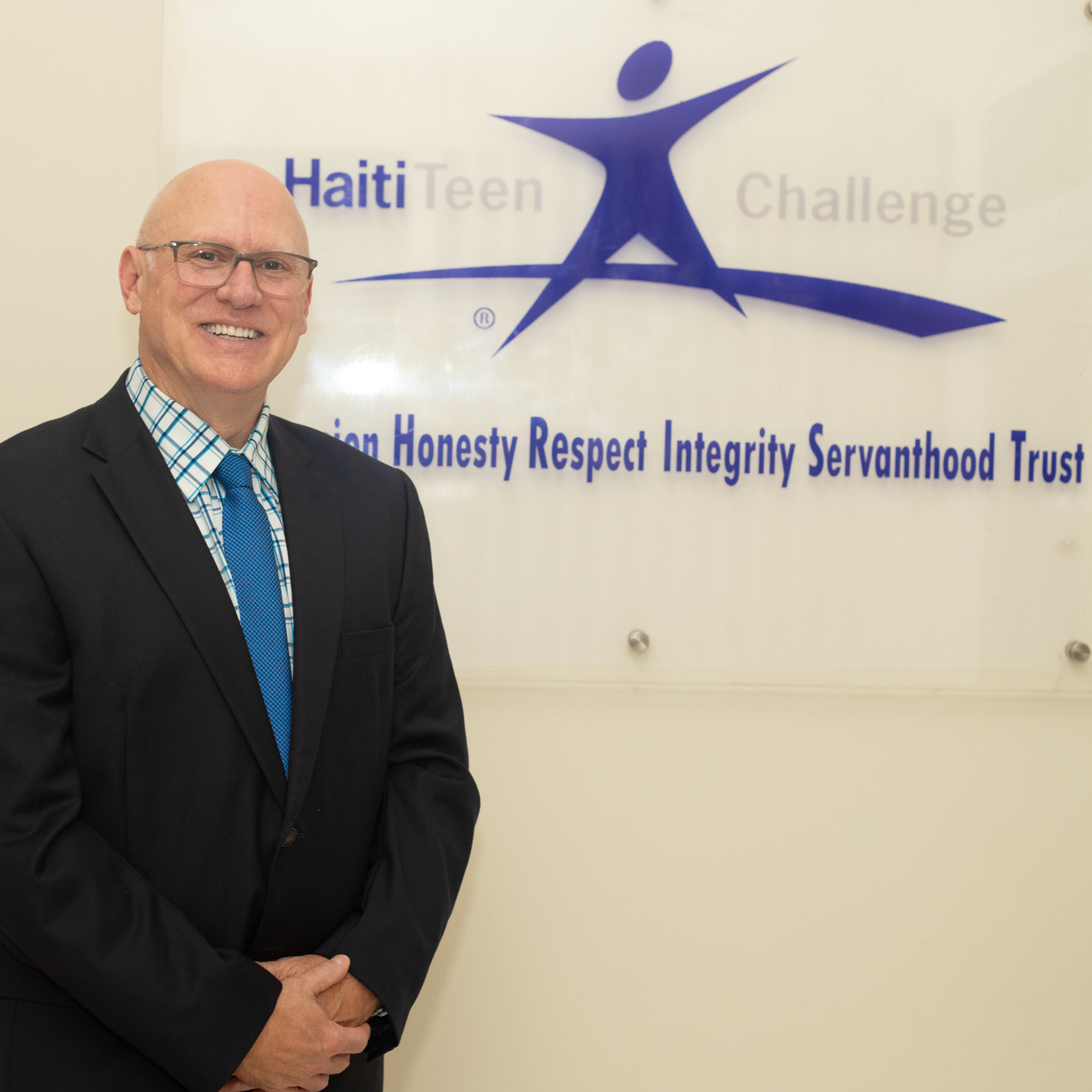 A smiling person in a suit and tie stands before a Haiti Teen Challenge sign listing core values.