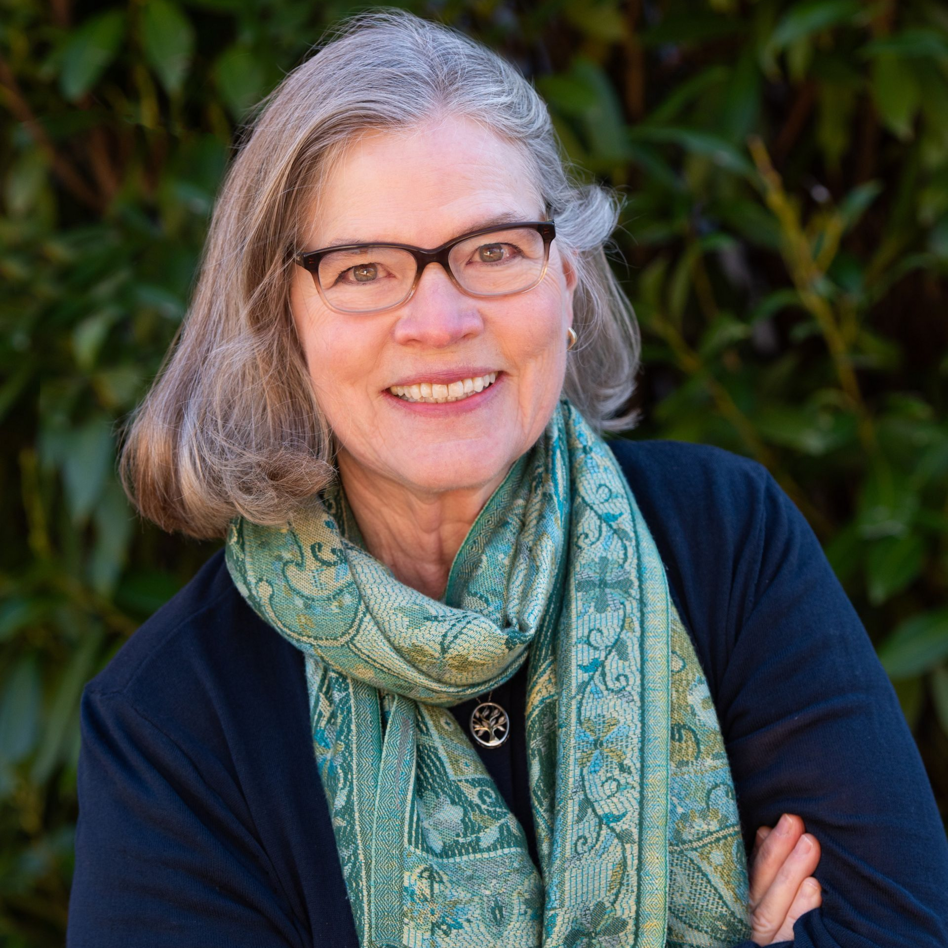 A smiling woman with shoulder-length grey hair and glasses, wearing a dark top and a green patterned scarf.