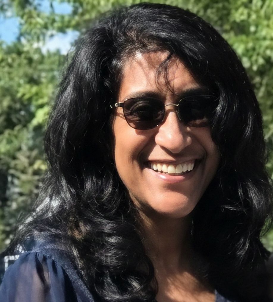 A smiling woman wearing dark-framed sunglasses and a dark blouse, set against a background of green foliage.