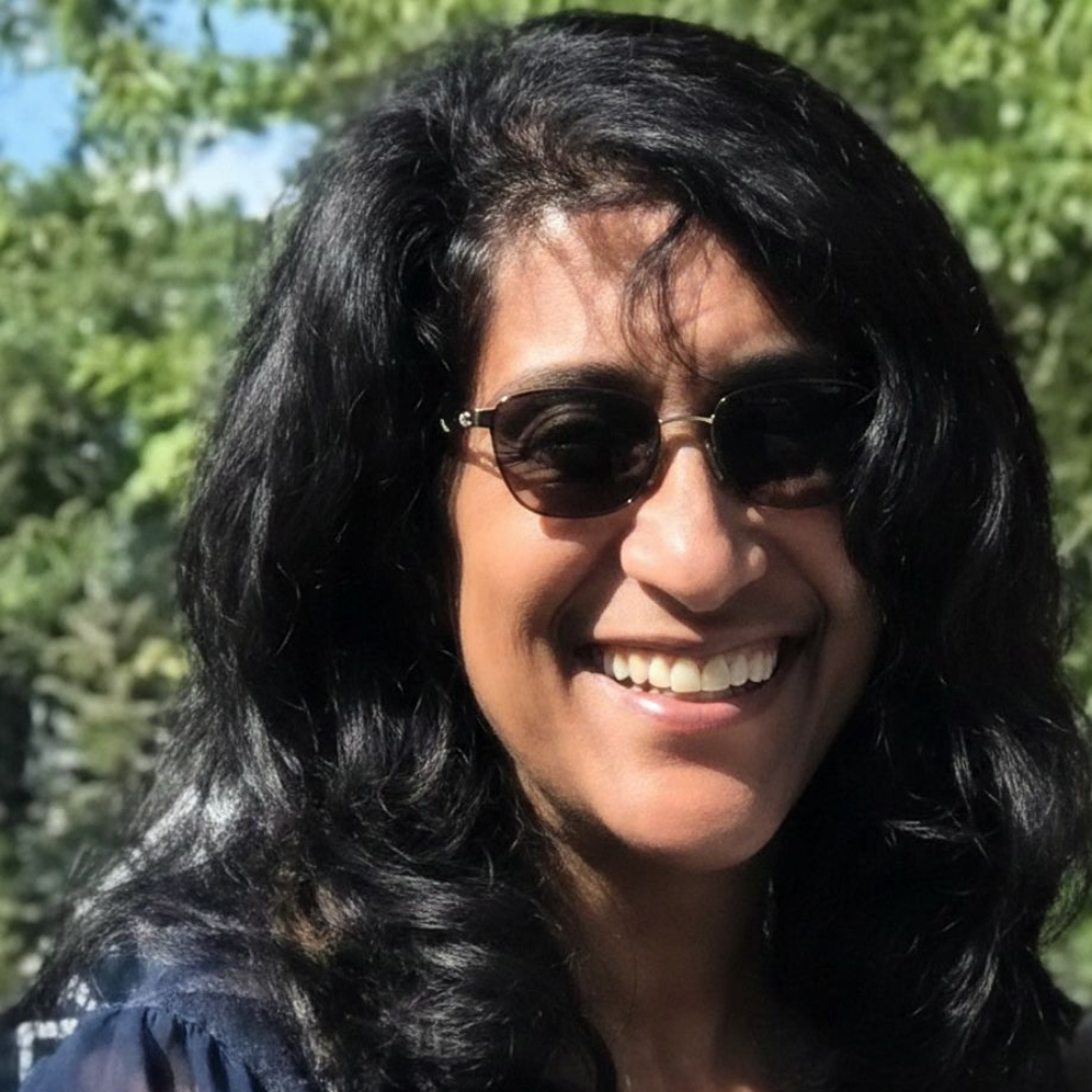 Woman with dark hair smiles, wearing sunglasses. Outdoors, blurred green background.