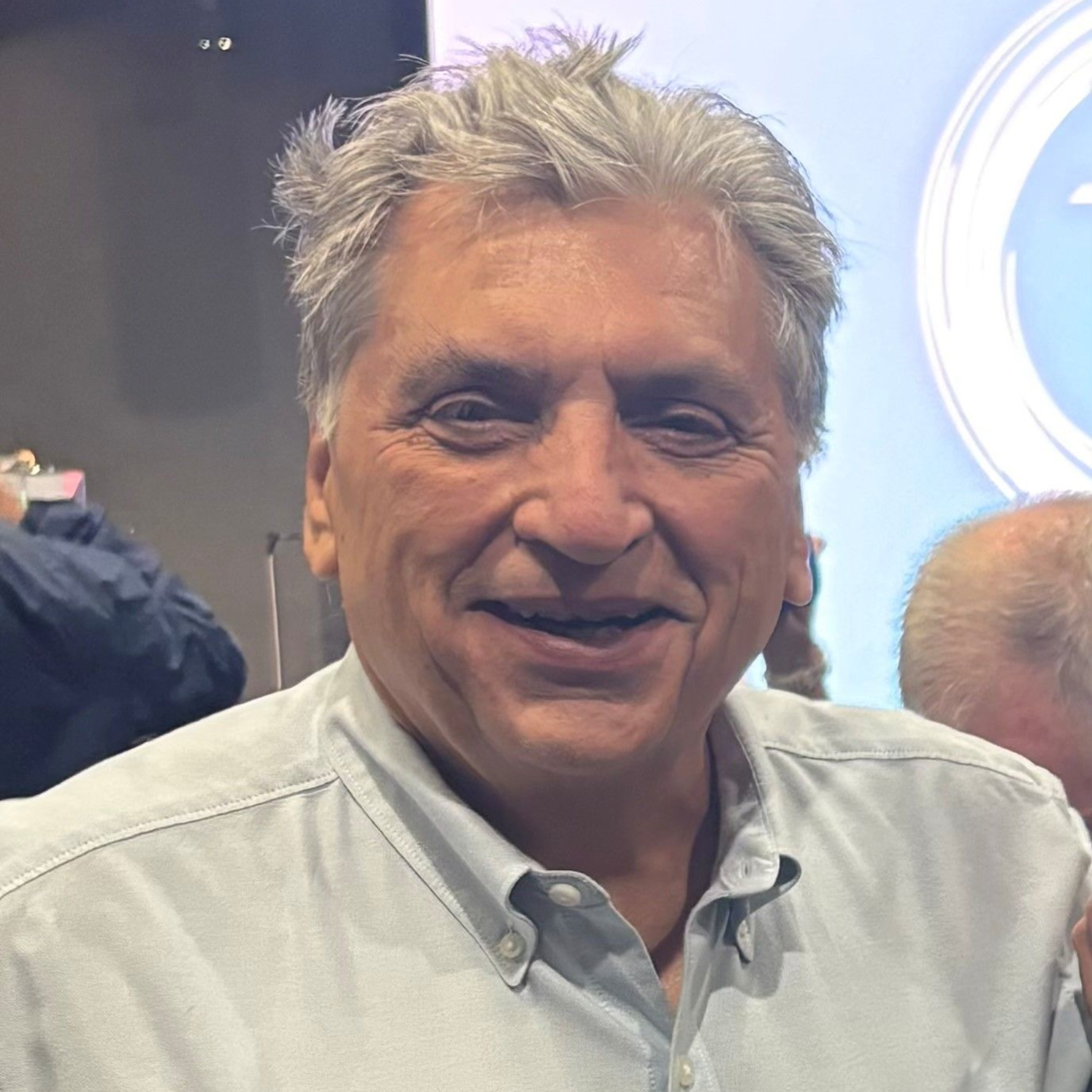 A person with light-colored, wavy hair and a light blue button-down shirt smiling in a brightly lit indoor setting.