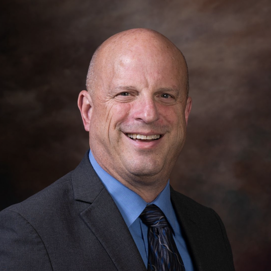 A professional headshot of a bald person wearing a dark grey suit, blue collared shirt, and a tie, smiling against a backdrop.