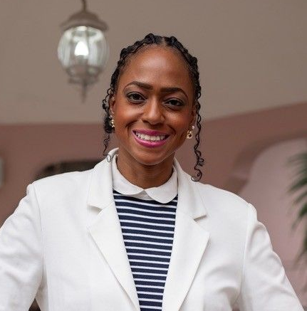 A smiling person with braided hair, wearing a white blazer over a navy and white striped collared shirt.